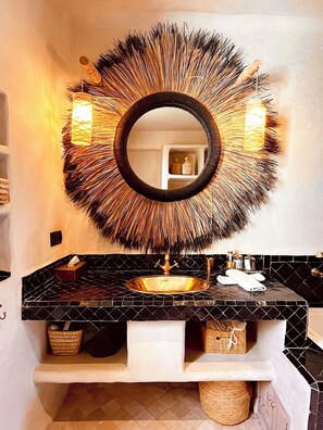 A bathroom with a round mirror, black and white tiled backsplash, and a gold sink. There are baskets on the counter and a towel rack. - Palais Bouchka (Essaouira)
