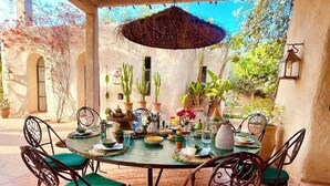 Mesa redonda com cadeiras estofadas em verde, um guarda-sol de palha e diversos pratos e bebidas.