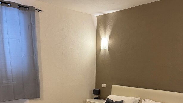 A neatly made bed with a dark bedspread and white towels, a bedside lamp, and a nightstand with a lamp. - Domu Grisìa Apartment Arzachena nice apartment in the center of the village (Arzachena)
