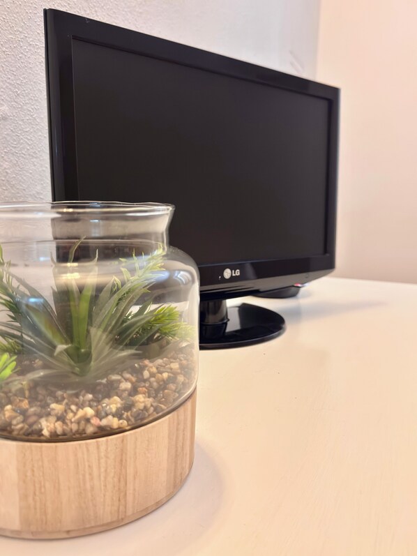 Ein klares Glas-Terrarium mit grünen Pflanzen auf einem Holzfuß, platziert auf einer weißen Oberfläche neben einem schwarzen Bildschirm.