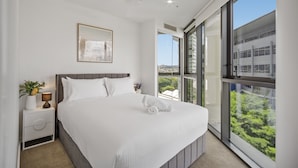 A modern bedroom with a large bed, a bedside table with a lamp, a framed picture on the wall, and a view of greenery through large windows. - Stylish 1-bed Apartment in South Brisbane (Brisbane)