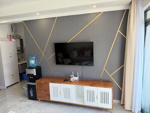 A modern living room with a flat-screen TV mounted on a grey wall with gold lines, a wooden console table with a water dispenser, and a white cabinet with a trash bin. - One bedroom in Lavington, Nairobi (Nairobi)