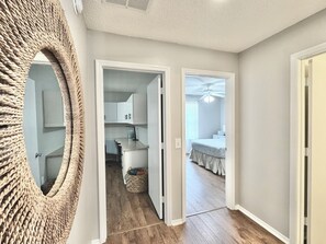 A hallway with a woven mirror, a kitchen with cabinets, and a bedroom with a bed and ceiling fan. - Casa Bella - 2/2 w/ Golf Cart & Grill in The Village of Summerhill (Summerhill, The Villages)