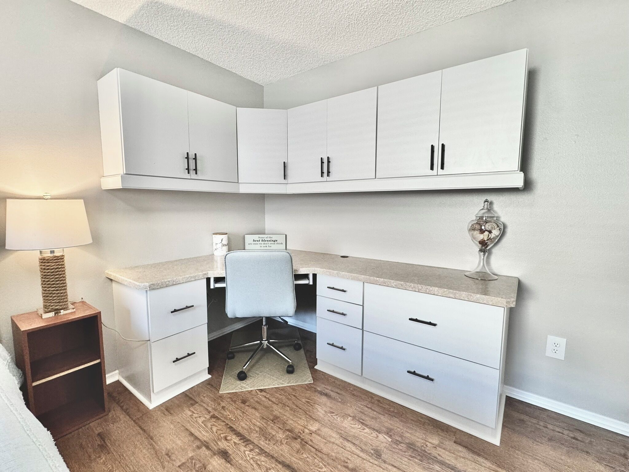 A corner bedroom with a desk, chair, and a bedside table with a lamp.