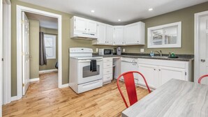 A compact kitchen with white cabinets, a microwave, an oven, and a dining area with a table and red chairs. - Peaceful Bangor Getaway | Clean & Cozy 2BR Home (Bangor)