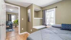 A bedroom with a bed, a bedside table, a window with curtains, a door leading to another room, and a small shelf. - Peaceful Bangor Getaway | Clean & Cozy 2BR Home (Bangor)