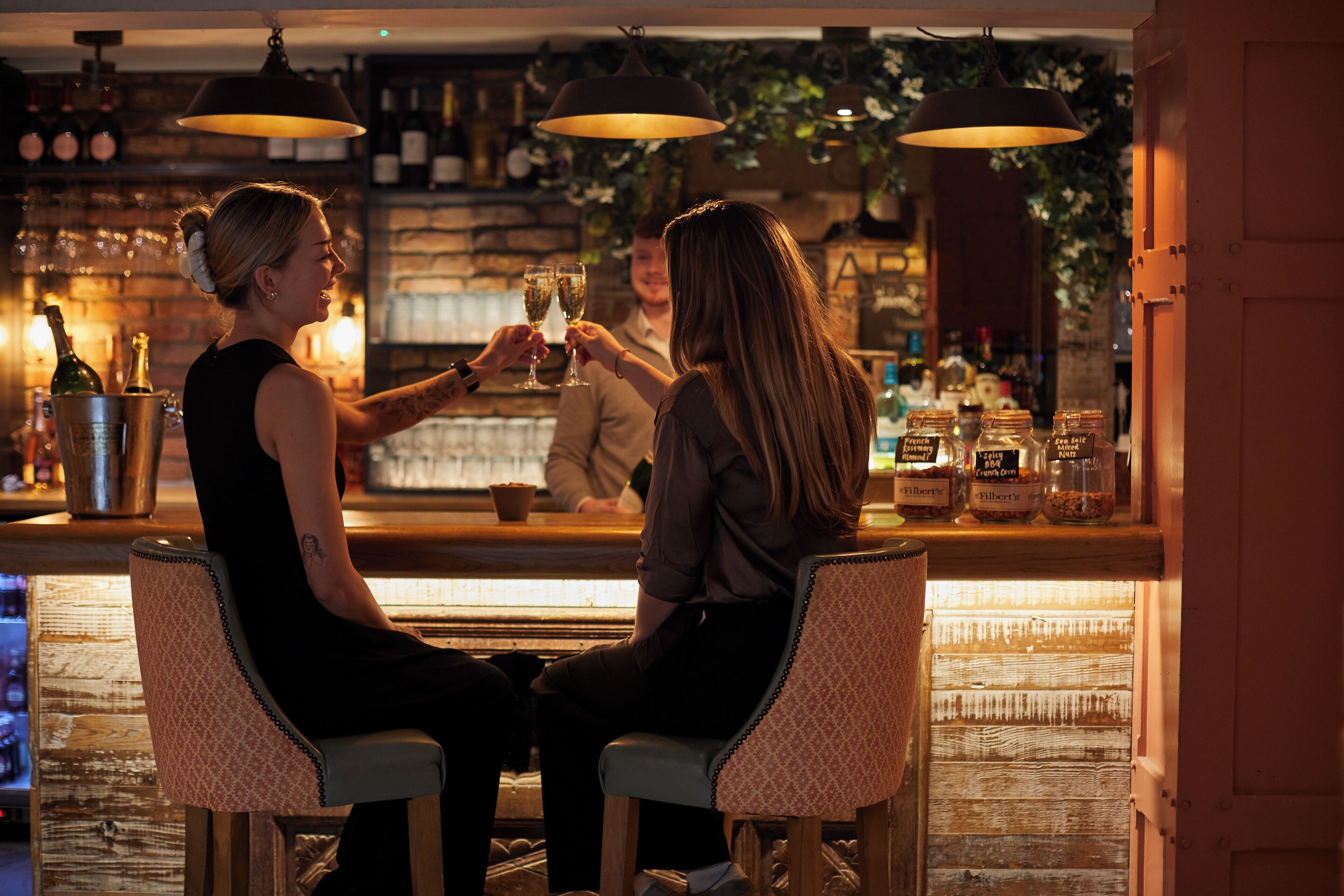 A bar area with two bar stools, a bartender, and a counter with bottles and glasses.