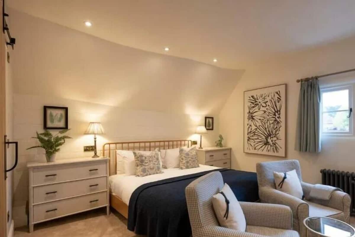 Un dormitorio con cama, mesitas de noche, cómoda, dos sillas, un cuadro y una ventana con cortinas.