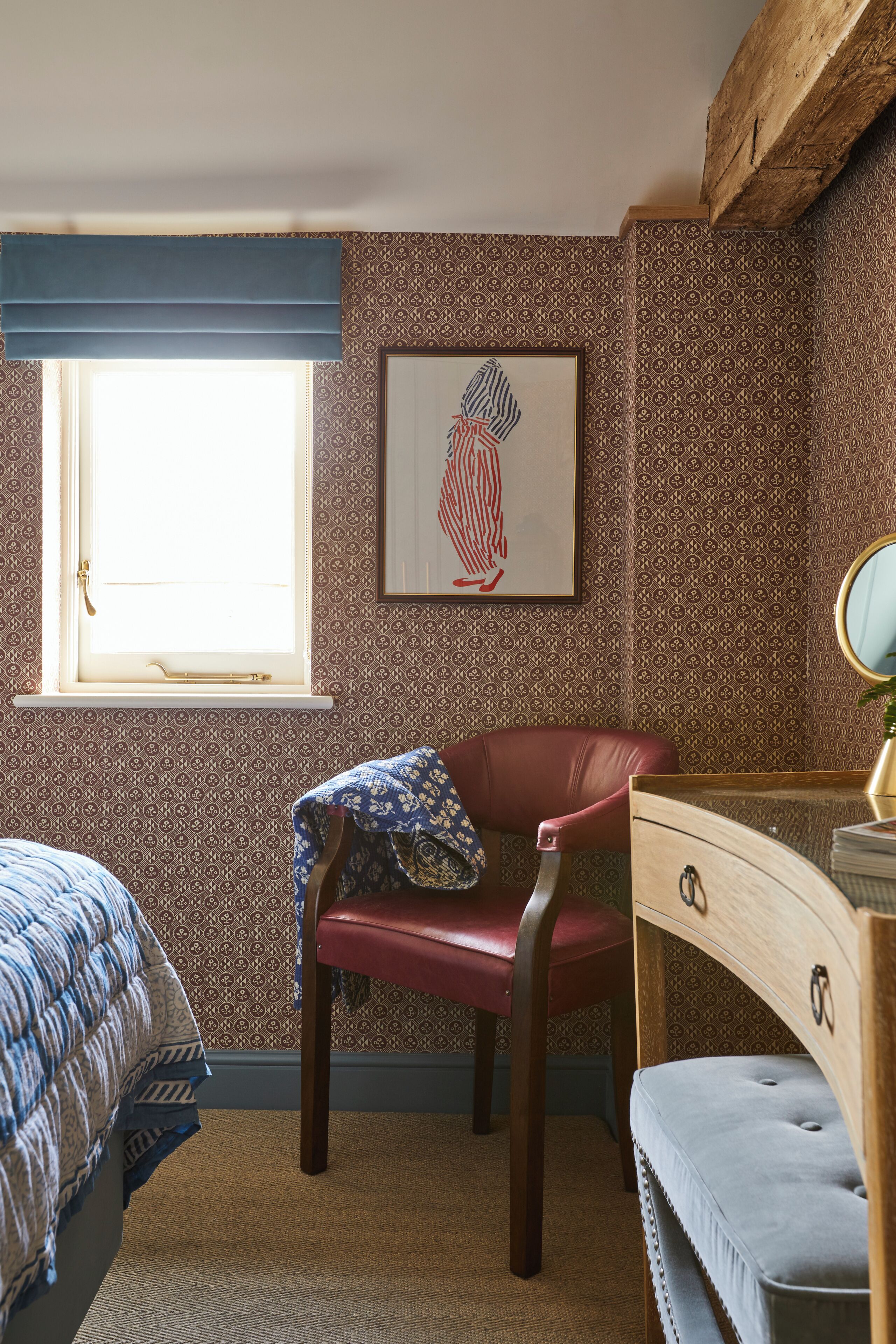 Ein Schlafzimmer mit Wandtapete mit Muster, einem Fenster mit blauem Vorhang, einem roten Sessel, einer Holzkommode und einem gerahmten Kunstwerk an der Wand.