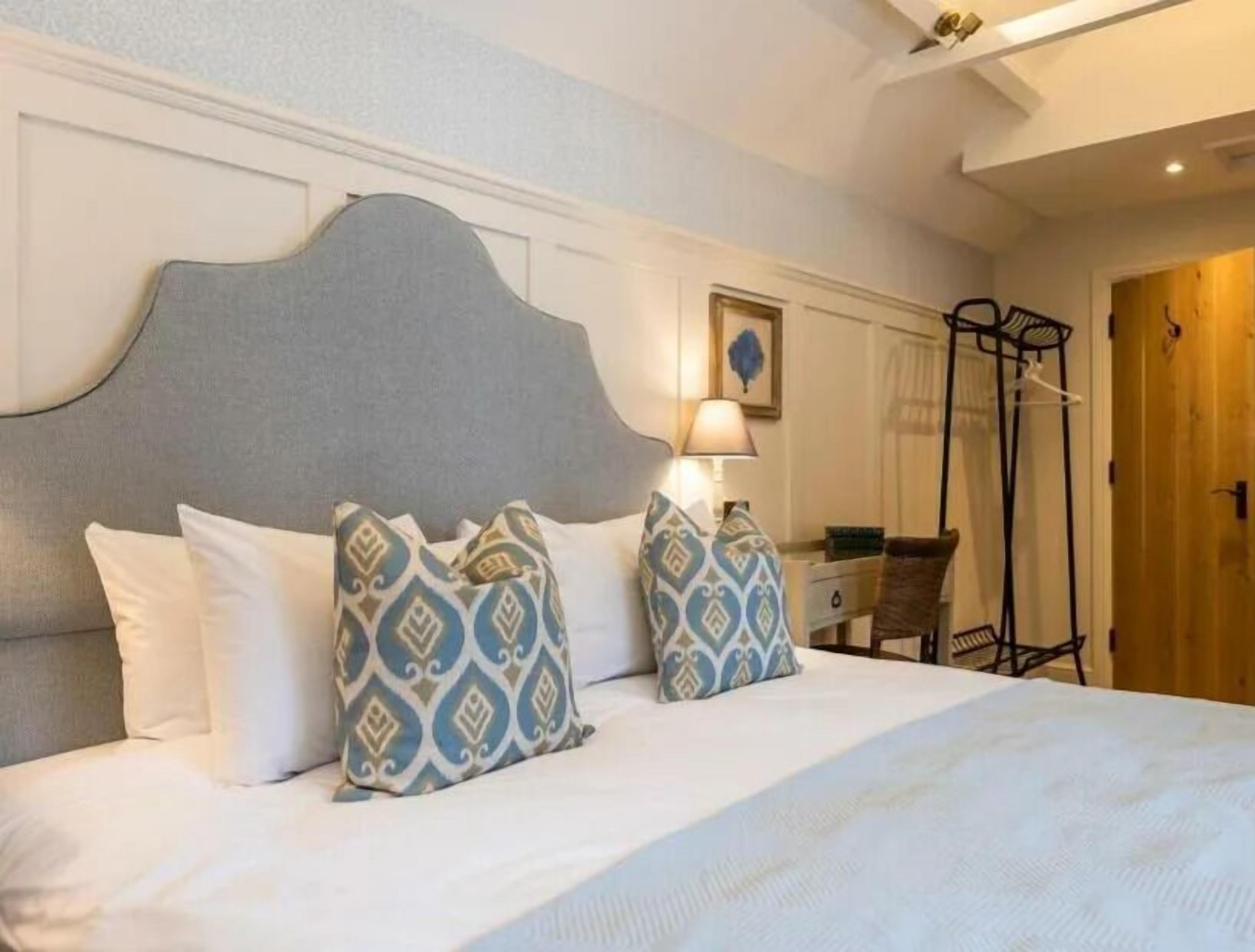 Una cama con almohadas blancas y estampadas, un cabecero, una mesita de noche con lámpara y una puerta de madera.