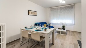 Un coin repas avec une table blanche dressée pour quatre personnes, un canapé bleu et une fenêtre ornée de rideaux à motifs.