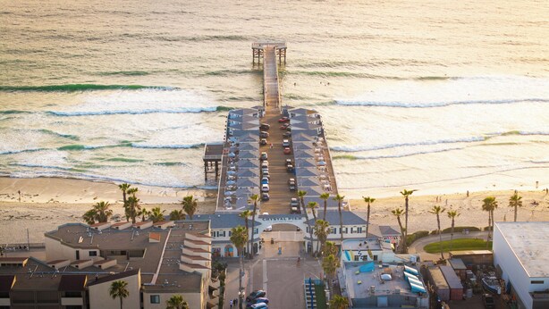 Een steiger die leidt naar een hotel met een parkeerplaats, strand en palmbomen.
