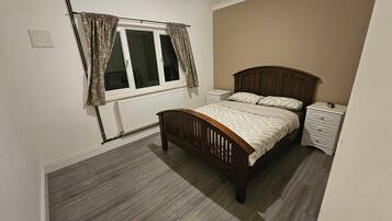 Een slaapkamer met een houten bed, een nachtkastje, een raam met gordijnen en een radiator.