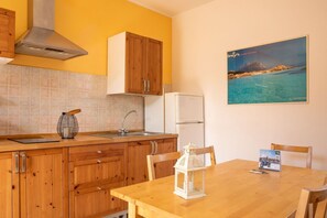 Eine Küche mit Holzausstattung, einer Edelstahl-Dunstabzugshaube, einem weißen Kühlschrank und einem Esstisch mit Stühlen. Ein gerahmtes Bild von einer Küstenlandschaft hängt an der Wand.
