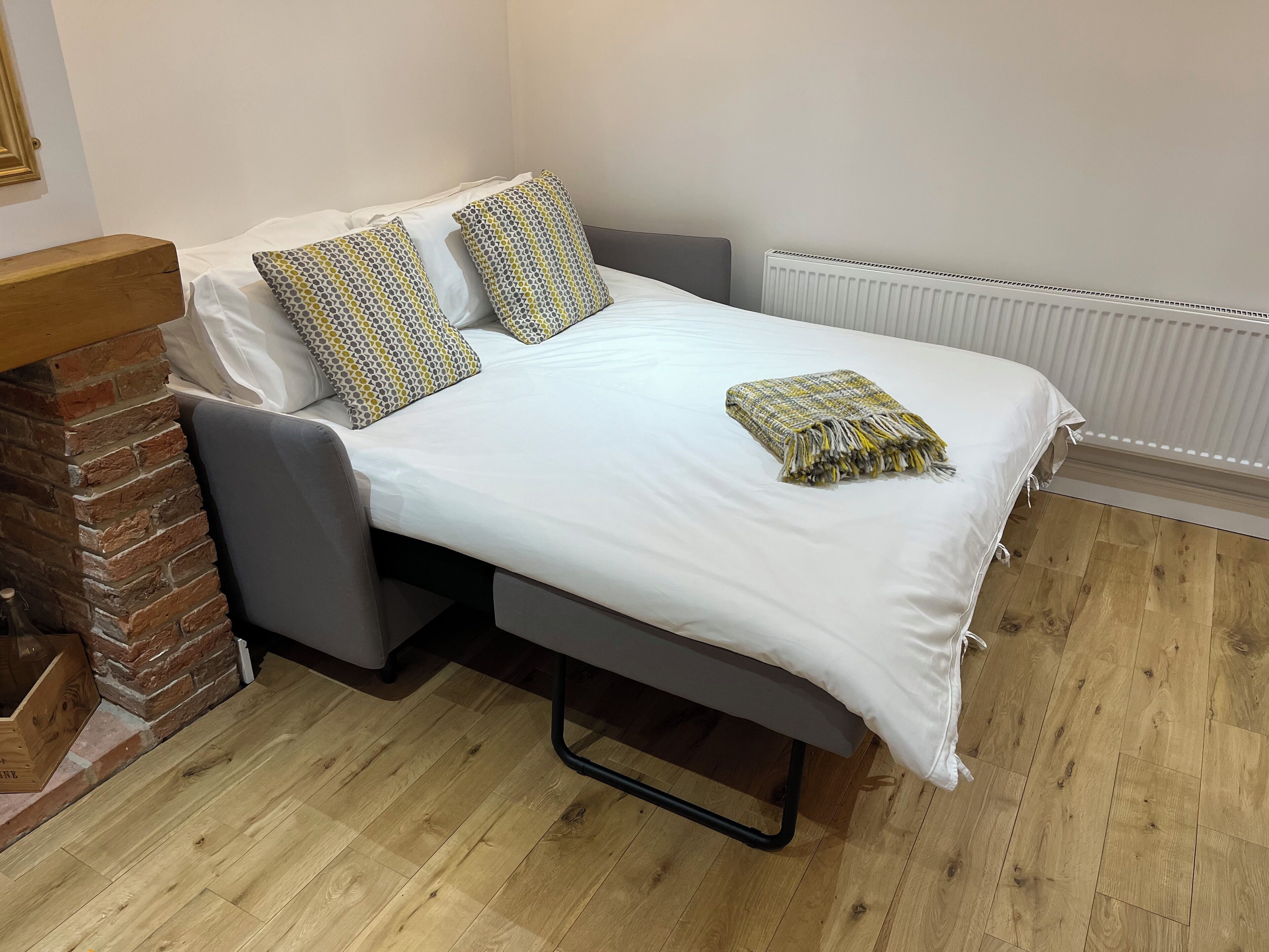 Habitación pequeña con una cama, dos almohadas estampadas, una manta, una pared de ladrillo, un radiador y suelo de madera.