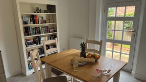 Een eetgedeelte met een houten tafel, witte stoelen, een boekenplank vol boeken, een schaal fruit en een raam met uitzicht op groen.
