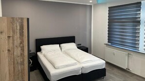 A modern bedroom with a black bed, white bedding, a wooden wardrobe, a nightstand, and a window with blinds. - Hotel Anker  (Linden)