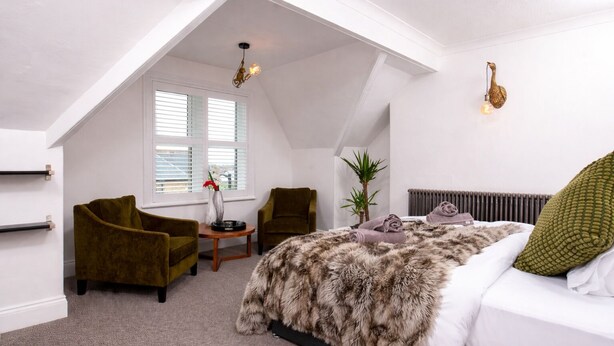 Een slaapkamer met een schuine kap, een bed met een zachte deken, twee fauteuils, een kleine tafel met een vaas en een radiator.
