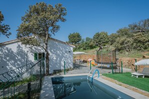 A swimming pool with a fence, a small building, and a grassy area with a lounge chair.