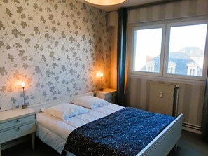 A bedroom with a bed, two bedside tables, a window with a view of buildings, and floral wallpaper. - THE NEST OF THE BACKFILL - Facing Grande Plage (Les Sables-d´Olonne)