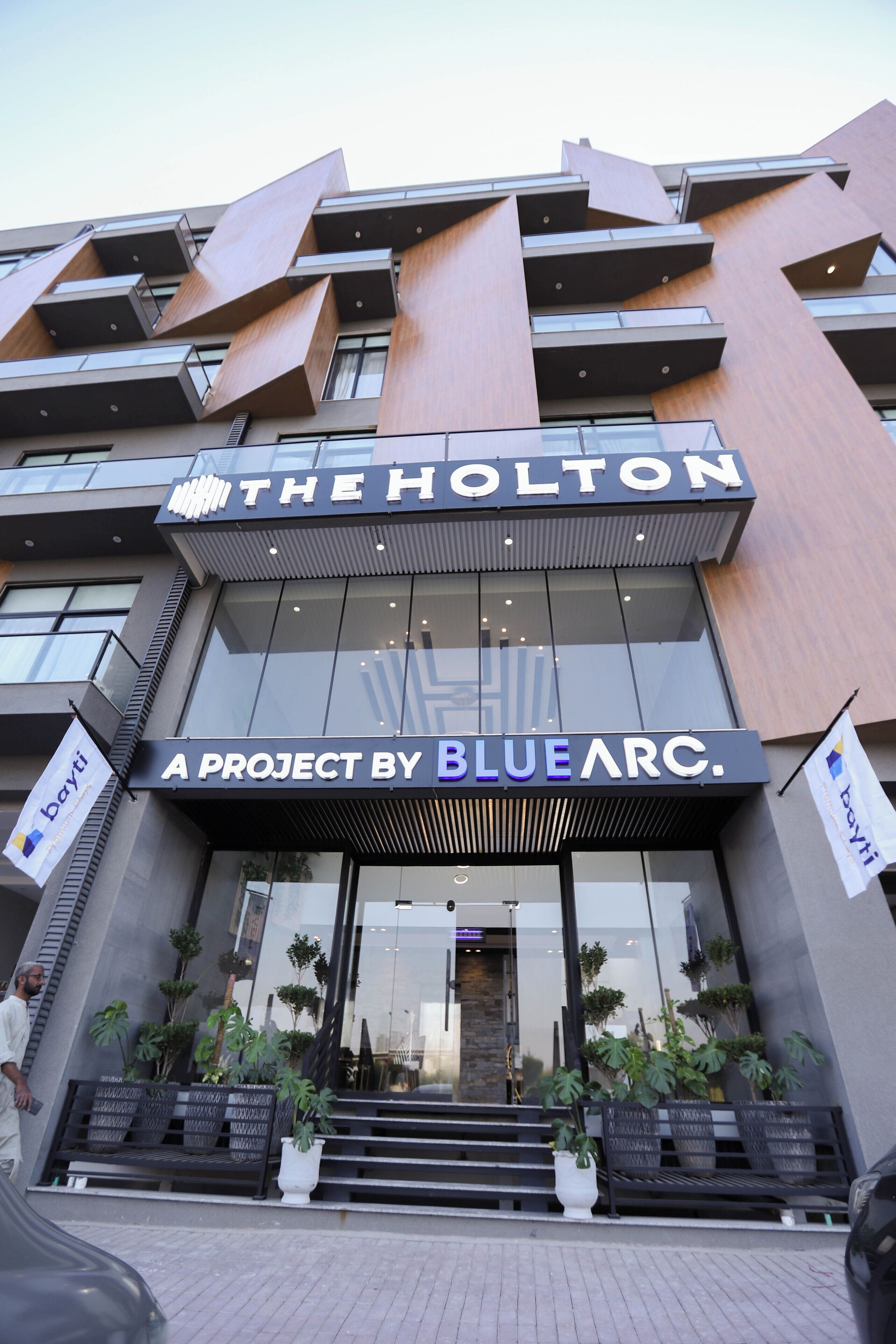 Un edificio moderno con il cartello "THE HOLTON A PROJECT BY BLUEARC". È caratterizzato da un ingresso ben visibile con gradini e piante in vaso.