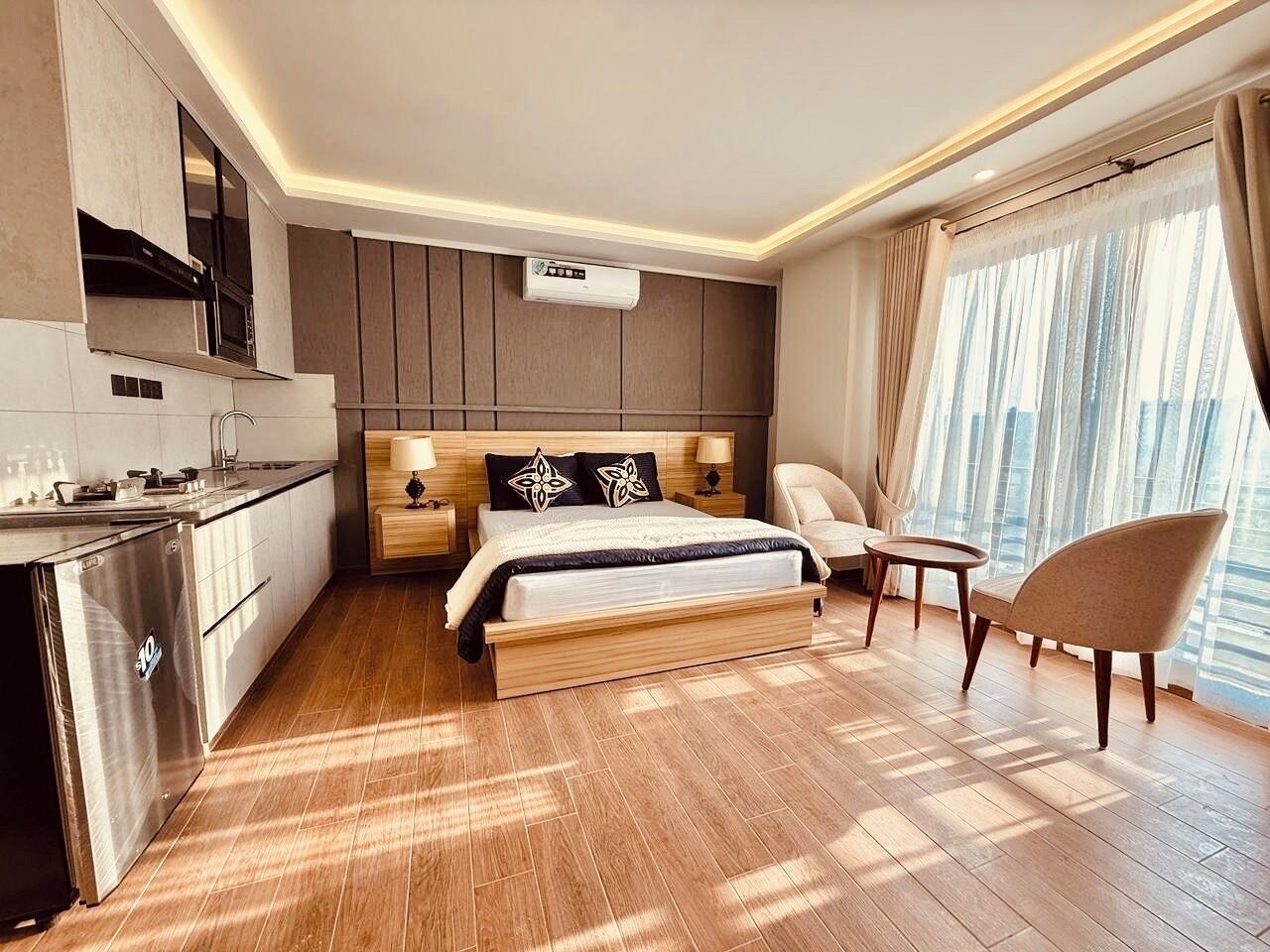A modern hotel room with a kitchenette, a bed with decorative pillows, a seating area, and large windows offering a city view.
