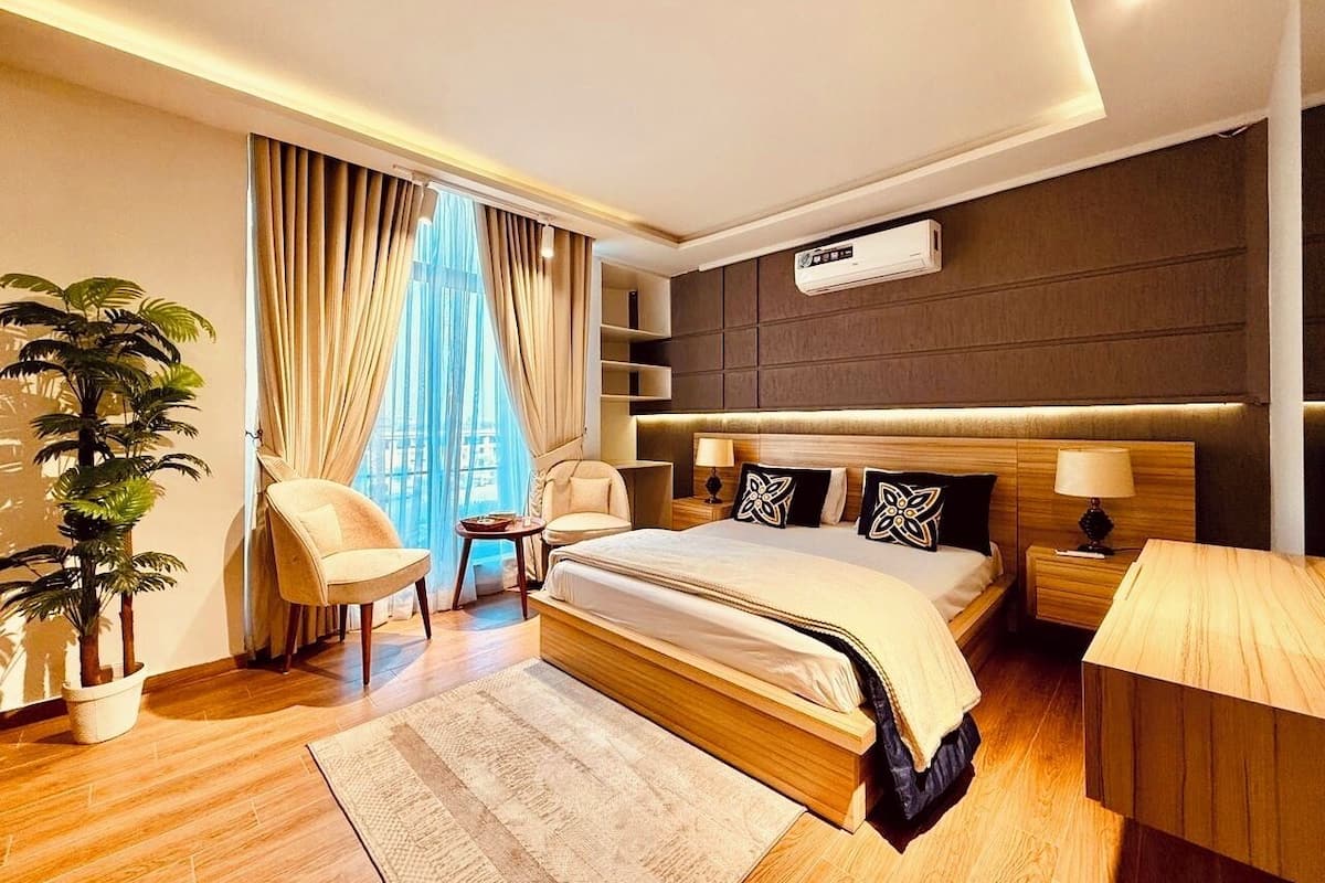 Ein modernes Hotelzimmer mit einem großen Bett, einem Nachttisch, einem Sessel, einer Topfpflanze und einem großen Fenster mit Vorhängen.
