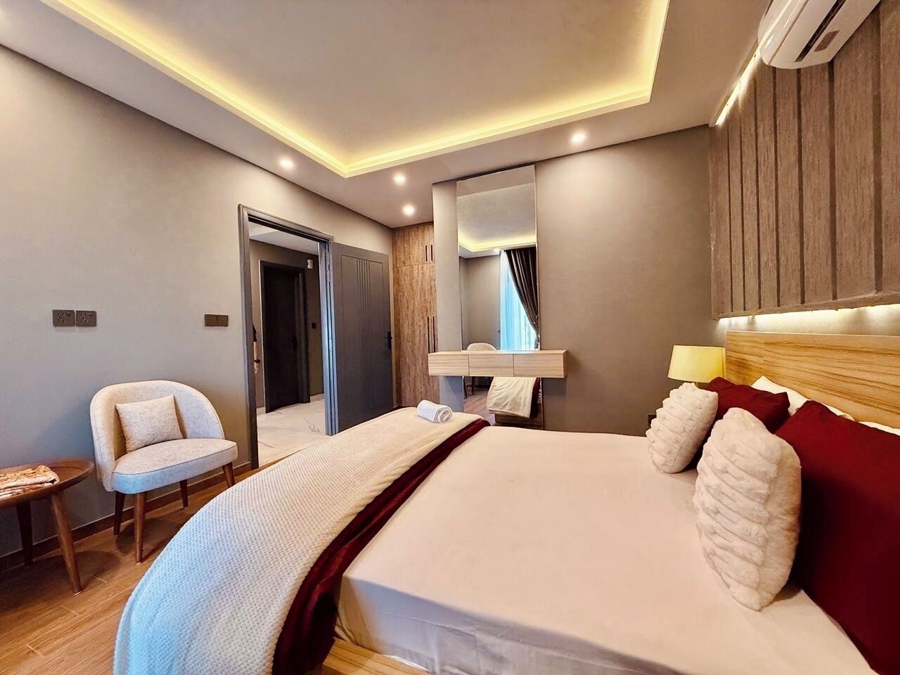 A modern hotel room with a large bed, a bedside table, a chair, a mirror, and a wooden headboard.