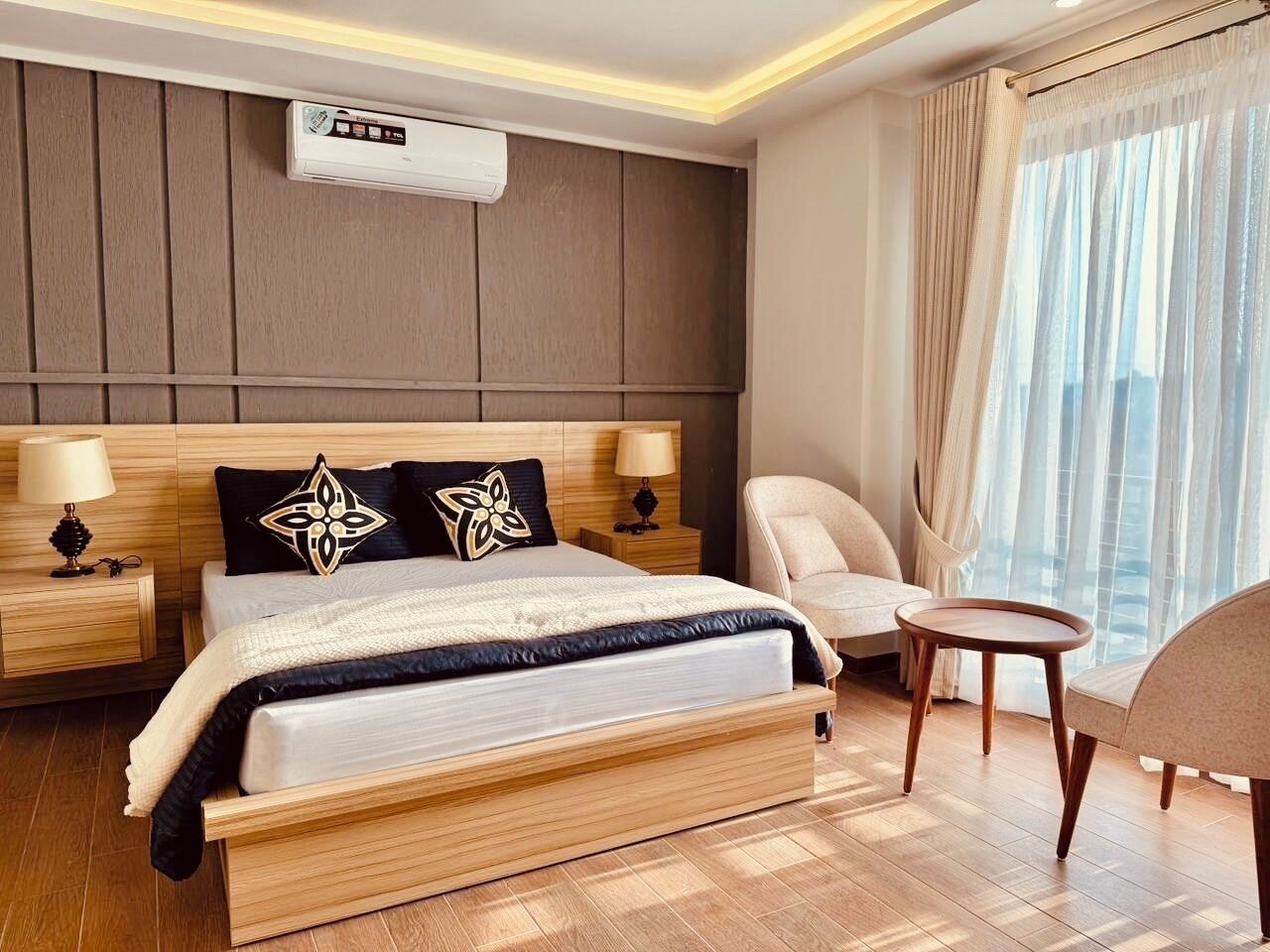 Una habitación de hotel moderna con una cama de madera, dos mesitas de noche con lámparas, una mesita redonda, una silla y un amplio ventanal con cortinas translúcidas.