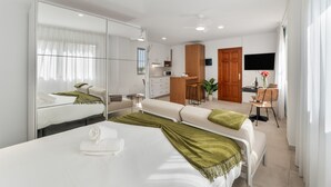 A modern hotel room with a large bed, a sitting area, a kitchenette, and a TV.