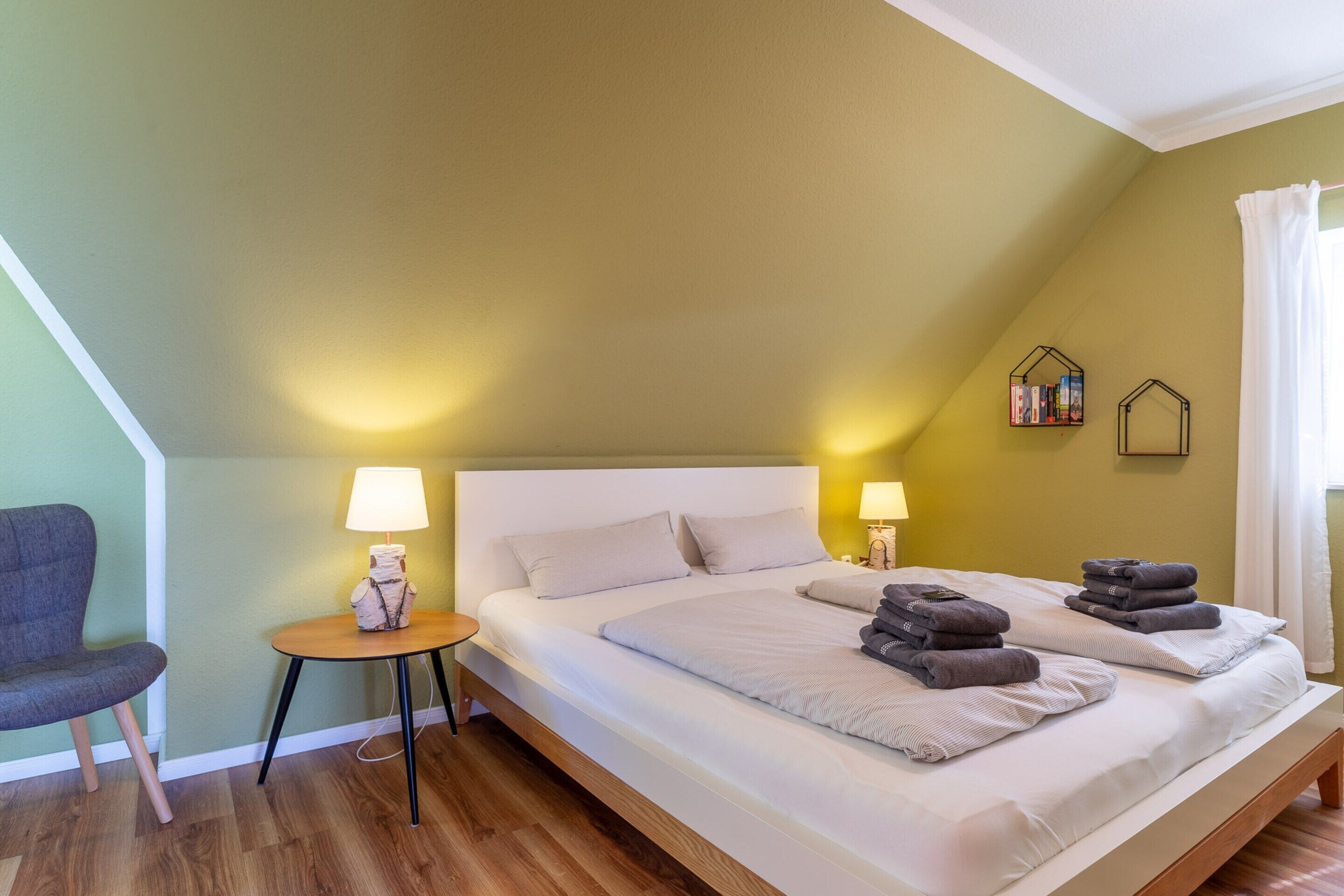Une chambre à coucher avec un lit simple, des lampes de chevet, une petite table, une chaise et une étagère remplie de livres.
