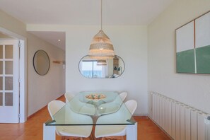 A dining area with a glass table and white chairs, a hanging woven light fixture, a round mirror, and a radiator on the wall. - Seafront Apt Torre Valentina (Calonge)