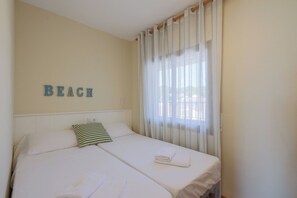 A bedroom with a bed, a pillow, a towel, a window with curtains, and a wall with the word "BEACH" spelled out. - Seafront Apt Torre Valentina (Calonge)