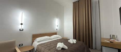 A hotel room with a bed, a bedside table, a chair, and a desk with a lamp.