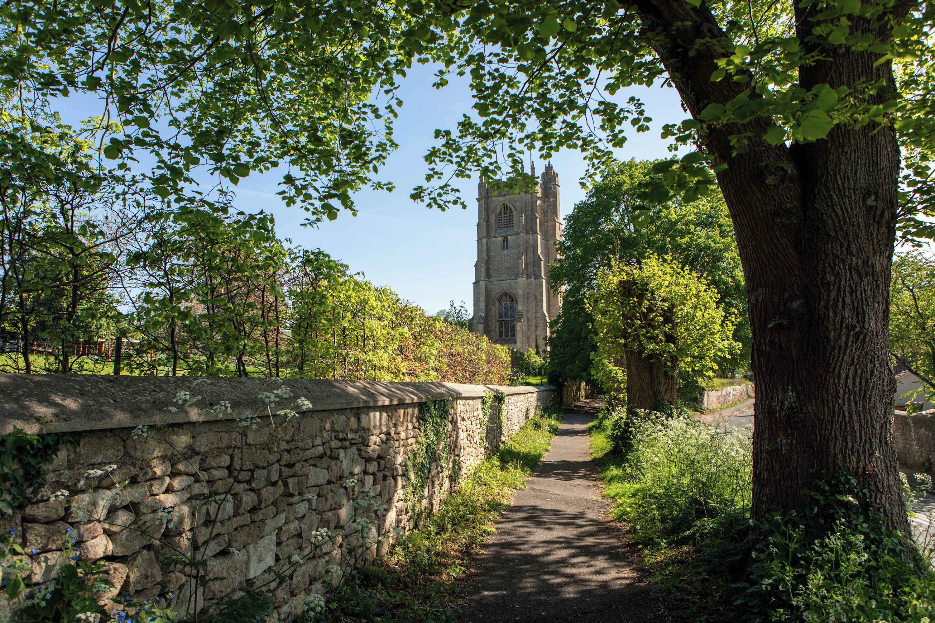 A stone pathway leading to a historic tower with a tall spire, surrounded by lush greenery and trees.