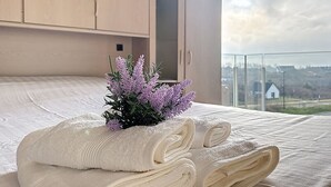 A neatly made bed with rolled towels and a small purple plant, a wooden wardrobe, and a view of a distant building through a glass door. - Flat Neptunus - Amazing seaview in De Panne (De Panne)