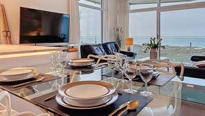 A modern hotel room with a dining area set for a meal, a living area with a sofa and a TV, and a large window offering a view of the sea. - Flat Neptunus - Amazing seaview in De Panne (De Panne)