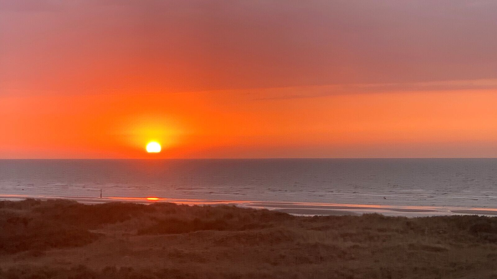 Un atardecer sobre una playa con el sol bajo en el horizonte, mar tranquilo y un primer plano con hierba.
