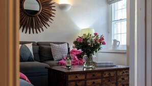 Ein gemütliches Wohnzimmer mit einer Couch, einem Holztisch mit Blumen in einer Vase, einem Wandspiegel und einem Fenster mit Aussicht.