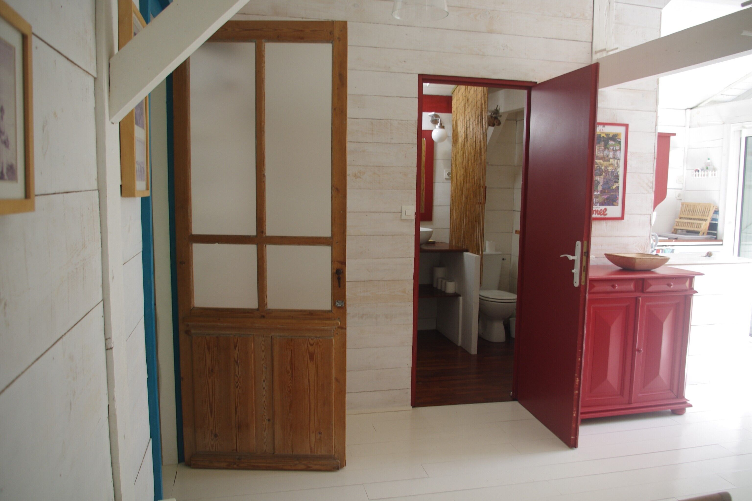Une pièce avec une porte en bois, un meuble rouge et une salle de bain visible à travers une porte ouverte.