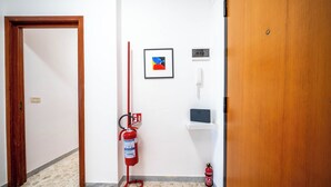 Een gang met een houten deur, een aan de muur bevestigde brandblusser, een ingelijst schilderij, een telefoon en een klein plankje.