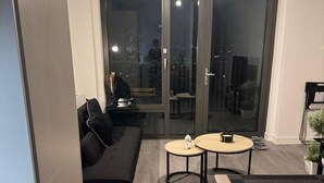 En moderne stue med en sort sofa, to runde borde og en skydedør, der fører ud til en udendørs balkon.