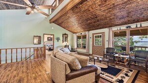 A spacious living area with a wooden ceiling, a large sofa, a dining table, and a view of the outdoors through large windows. - Scenic Serenity | Maggie Valley, 4BR, 3 BA, VIEWS! (Maggie Valley)
