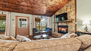 A cozy living room with a stone fireplace, wooden ceiling, and a view of the outdoors. - Scenic Serenity | Maggie Valley, 4BR, 3 BA, VIEWS! (Maggie Valley)