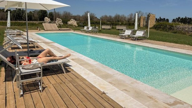 Un espace aménagé au bord de la piscine, avec des chaises longues et des parasols, une piscine à l’eau d’un bleu limpide et un mur de pierre en arrière-plan.