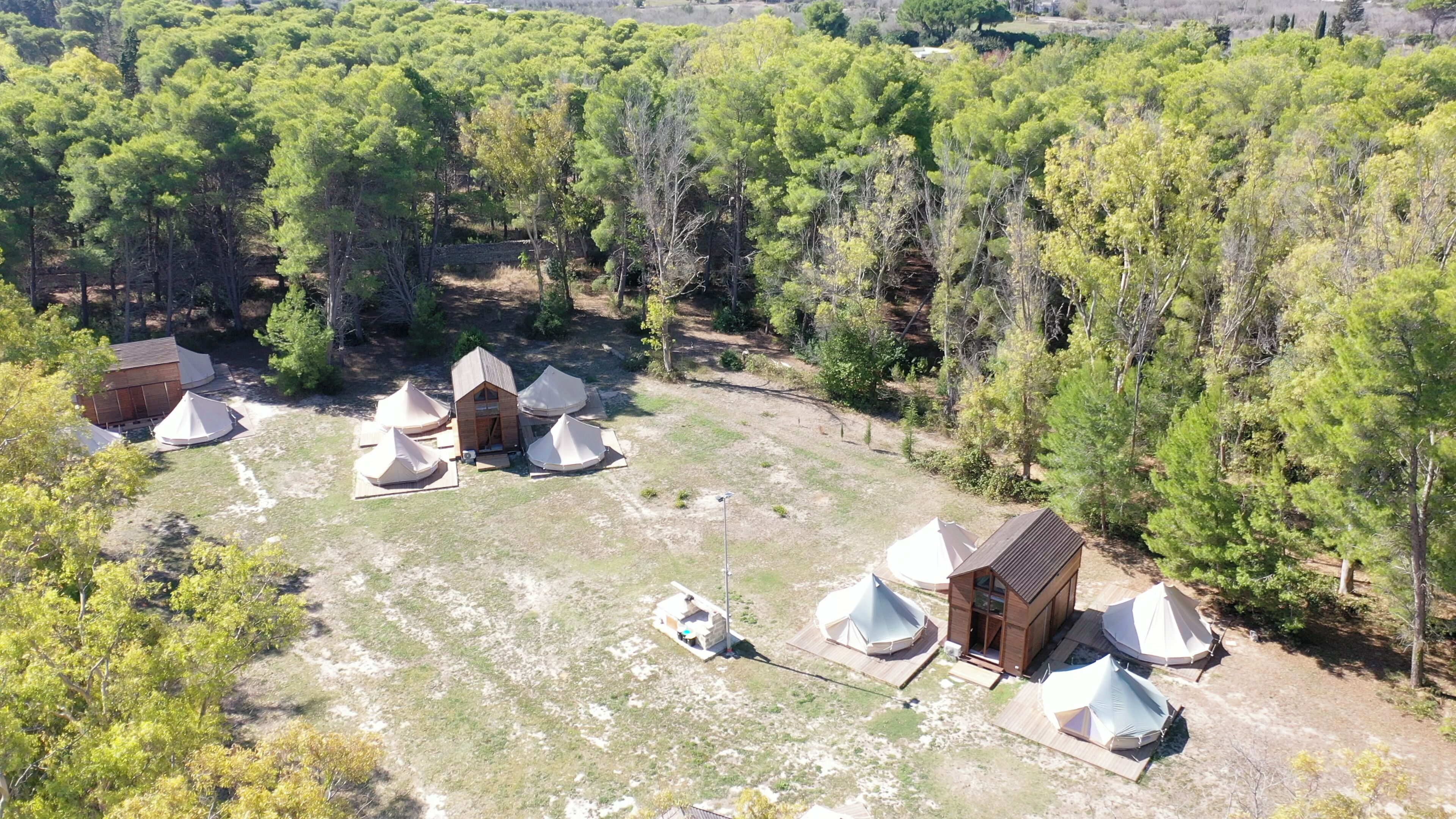 An aerial view of a campsite with several tents and wooden cabins surrounded by dense forest.