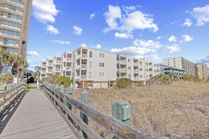A wooden boardwalk leading to a beachfront residential area with multi-story buildings, balconies, and a clear sky. - Ocean Pier Unit 303 Sleeps 6-2br/2ba-Full Kitchen-Pool-Direct Oceanfront Balcony (North Myrtle Beach)
