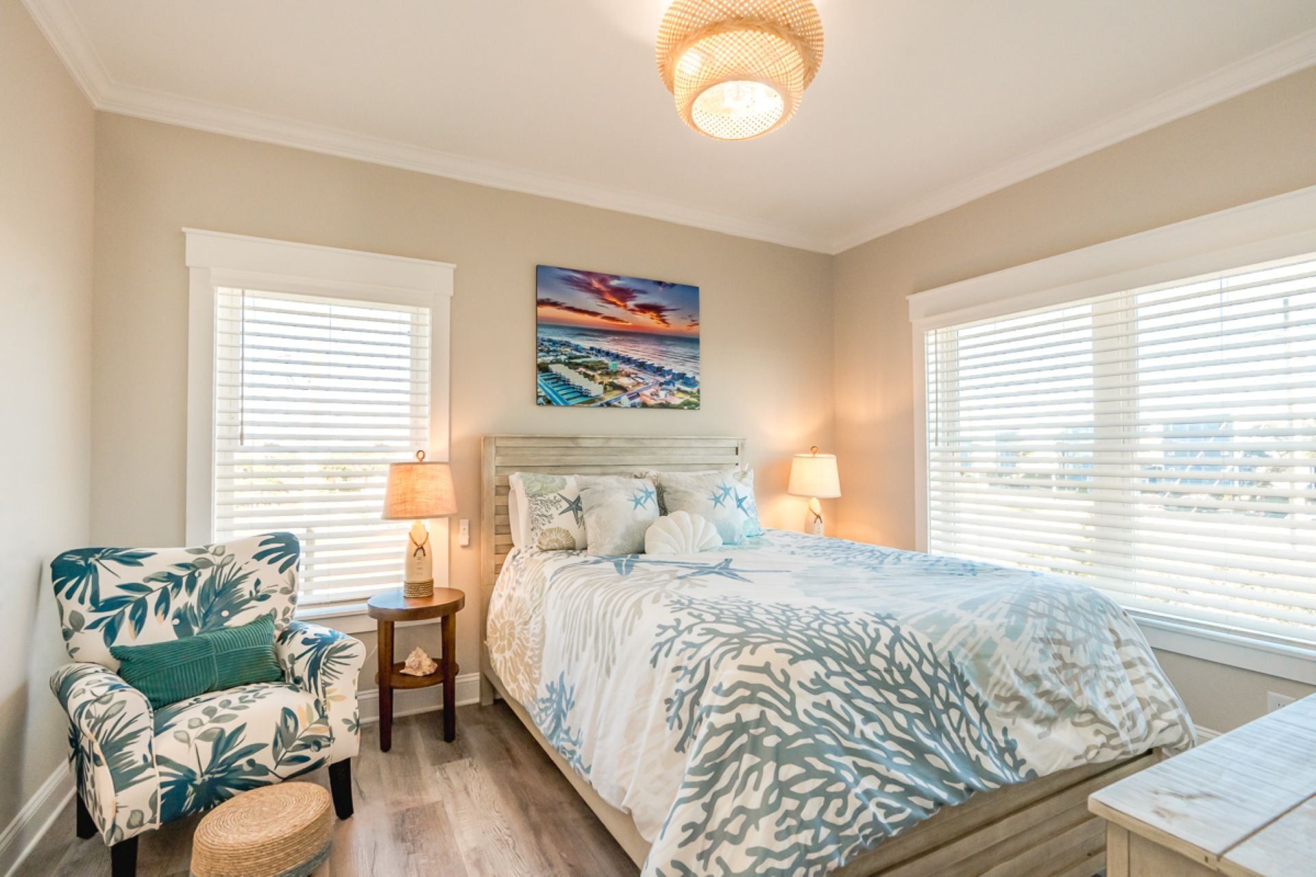 A bedroom with a bed, two bedside lamps, a chair with a tropical print, a painting on the wall, and a window with blinds.