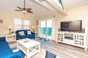 A living room with a sofa, coffee table, TV, and a balcony with chairs. - A Porpoiseful Life - Oceanfront 4 BR Duplex, 2 Decks and Dog Friendly! (N. Topsail Beach)