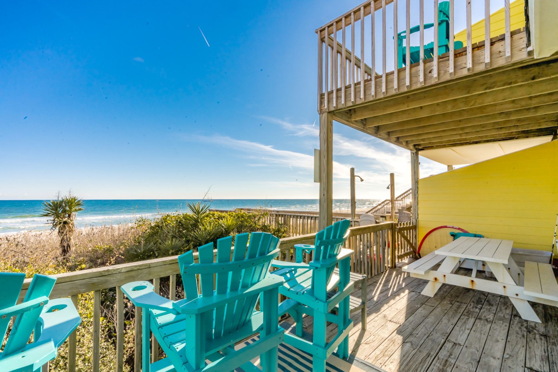 Een houten terras met turquoise stoelen en een picknicktafel met uitzicht op een strand.